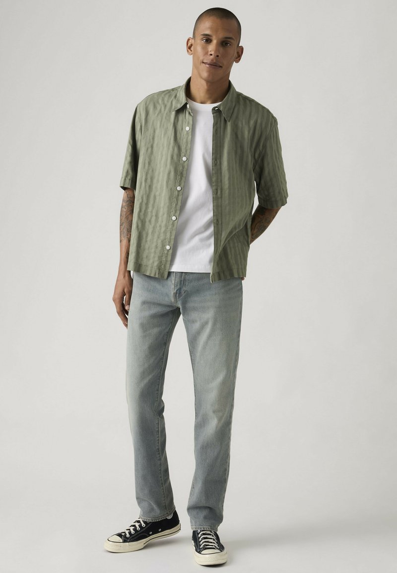 Homme aux cheveux courts portant une chemise à manches courtes vert clair boutonnée, un T-shirt blanc, un jean bleu clair et des baskets noires, debout de manière détendue.