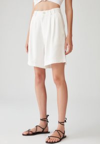 Pantalones cortos de lino blancos con un corte relajado, frente con pliegues y cierre de botón. Combinados con sandalias negras con tiras. Textura suave y diseño nítido.