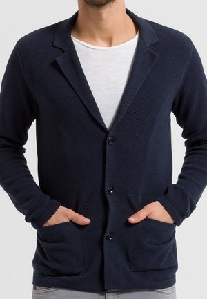 Uomo che indossa un cardigan blu navy con bottoni sopra una maglietta bianca, con le mani nelle tasche anteriori, su uno sfondo neutro.