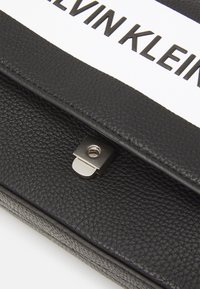 Černá kožená taška s texturovaným povrchem, s kovovým stříbrným zapínáním a bílou etiketou s nápisem "KELVIN KLEIN" tučným písmem.