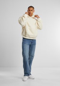 Cremefarbene, fuzzy Kapuzenjacke mit einer Fronttasche und Kordelzugkapuze, kombiniert mit hellblauen Jeans und weißen Sneakers. Glatte Textur.