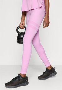 Ροζ κολάν με λεία υφή και κατακόρυφες ρίγες στους μηρούς, σε συνδυασμό με μαύρα αθλητικά παπούτσια με σχεδίαση από δίχτυ και στρογγυλή σόλα.