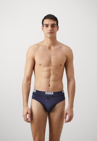 Tamno modre hlače z belim pasom, ki se kontrastno izstopa in nosi logo "PUMA". Gladka tekstura in prilegajoč dizajn.