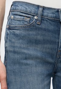 Κοντινό στους γοφούς και την τσέπη ενός μπλε τζιν με ορατά ραφές και μεταλλικά καρφιά, φορεμένο από ένα άτομο με το χέρι ακουμπισμένο στο πλάι.