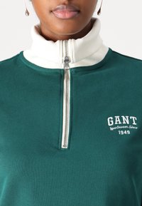 Sudadera verde con cierre y cuello de canalé blanco. Presenta un zipper plateado y un logo bordado que dice "GANT Sportswear Since 1949". Textura suave.