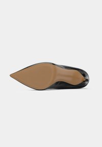 Escarpin noir en vernis, à bout pointu, vu de la semelle avec un dessous texturé marron et un talon étroit, sur fond blanc.