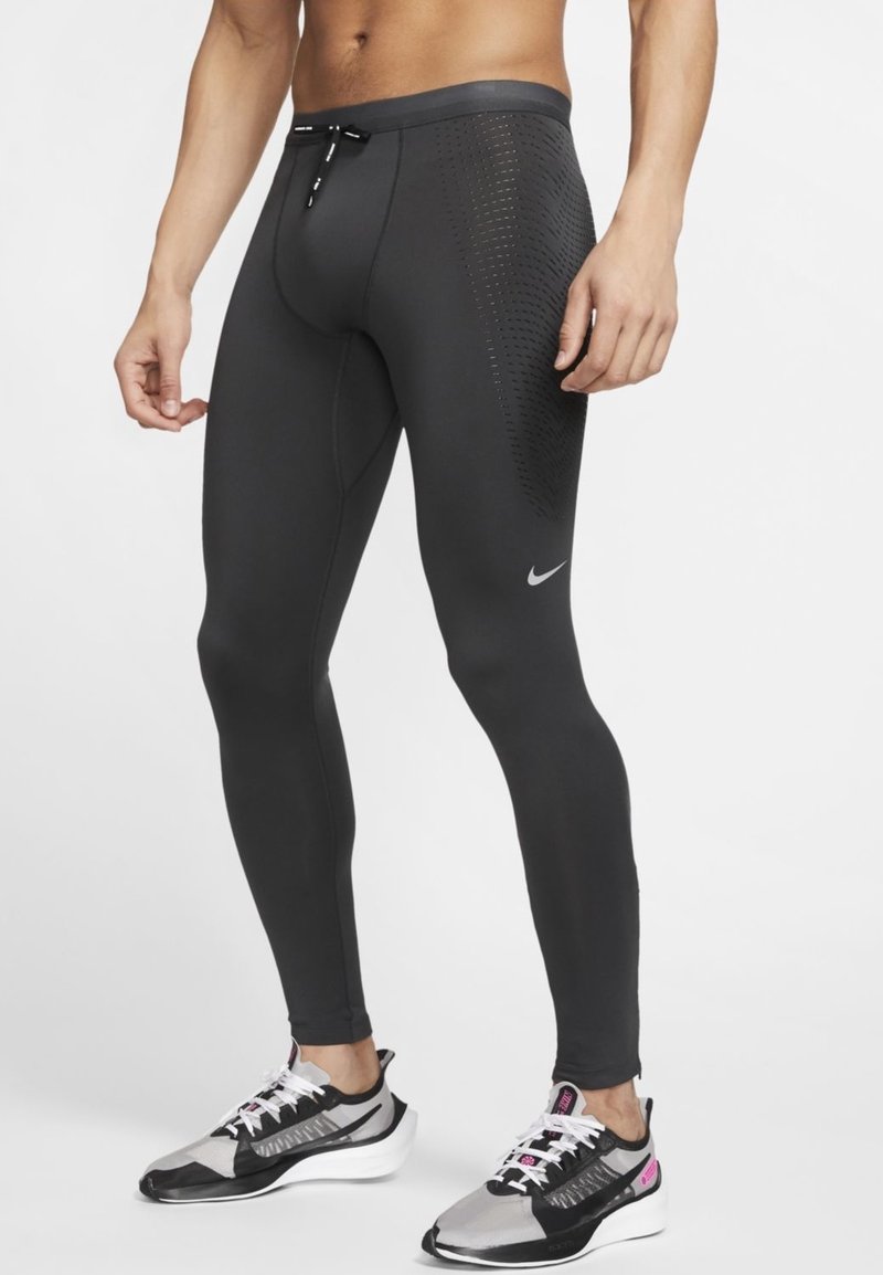 Męski model ubrany w czarne legginsy kompresyjne Nike z bocznymi panelami wentylacyjnymi oraz szare, czarne i białe buty biegowe Nike z różowymi akcentami.