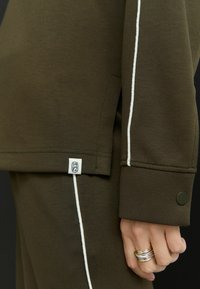 Sweatshirt vert olive avec des passepoils blancs le long des coutures. Étiquette de logo notable sur l'ourlet, tissu mélangé en coton et détail de bouton aux poignets.