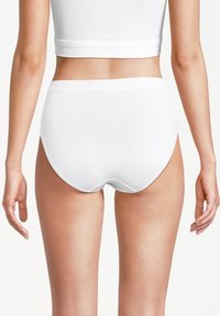 Frau in weißen, hochtaillierten Baumwollunterhosen und einem passenden Cropped-Top, von hinten vor einem einfarbigen Hintergrund gezeigt.
