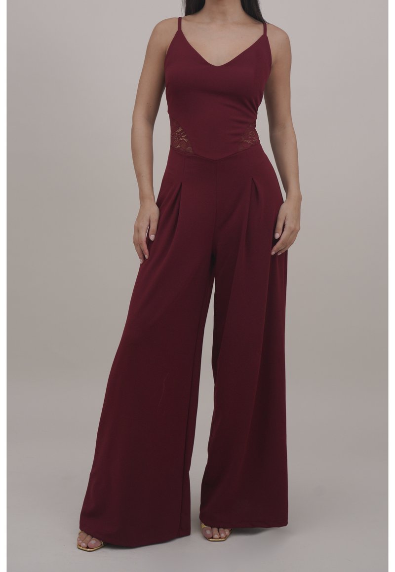 Femme portant une combinaison bordeaux sans manches avec des panneaux latéraux en dentelle et un pantalon à jambes larges, debout devant un fond uni.