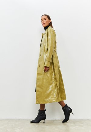 Trench coat lungo color kaki con finitura lucida, design doppiopetto e vita con cintura. Indossato con stivali neri con punta a punta.