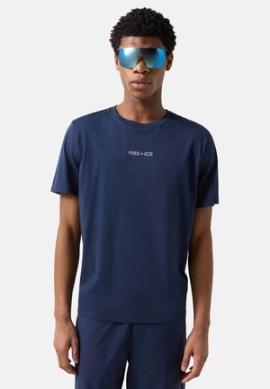 Junger Mann trägt ein marineblaues "FIRE+ICE" T-Shirt und passende Hosen mit großen reflektierenden blauen Sonnenbrillen vor einfachem Hintergrund.