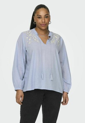 Blouse bleu clair à manches longues, décolleté en V, avec des broderies florales sur la poitrine et un cordon de serrage avec des pompons. Tissu en coton.