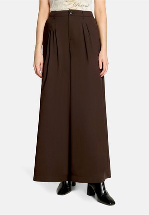 Femme portant un pantalon marron taille haute à jambes larges avec plis devant, associé à des bottines noires à talons et bout carré.