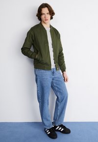 Veste bomber vert olive avec une fermeture éclair, chemise grise en dessous, jeans bleus et baskets noires avec des rayures blanches ; pose debout contre un fond plain.
