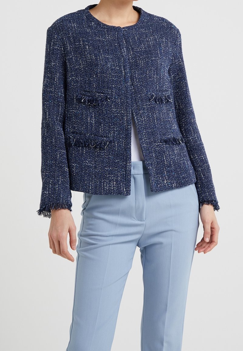 Meren värinen tweed-takki, jossa on pyöreä kaulus, avoin etuosa ja frillitykset taskuissa ja hihansuissa, yhdistettynä vaaleansinsisiin housuihin.