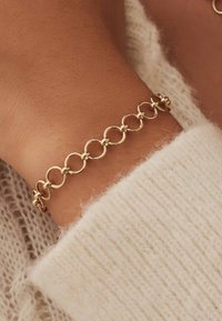 Goldene Kettenarmband mit runden Gliedern und glatten Oberflächen. Trägt ein Handgelenk, das teilweise von einem strukturierten, cremefarbenen Pullover bedeckt ist.