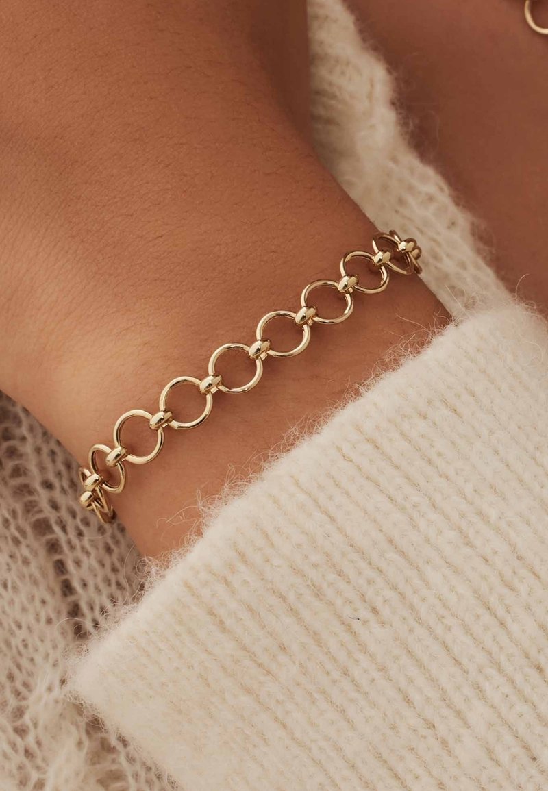 Goldene Kettenarmband mit runden Gliedern und glatten Oberflächen. Trägt ein Handgelenk, das teilweise von einem strukturierten, cremefarbenen Pullover bedeckt ist.