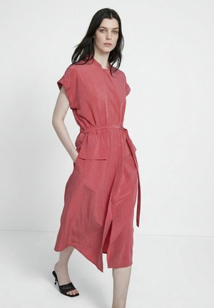 Kobieta o ciemnych włosach, ubrana w koralową sukienkę midi z paskiem i kieszeniami, nosząca czarne sandały na obcasie z odkrytymi palcami, stojąca na białym tle.