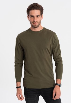 Olijfgroene t-shirt met lange mouwen van katoen. Heeft een ronde halslijn en aansluitende mouwen, gedragen met een donkere jeans en een polshorloge.