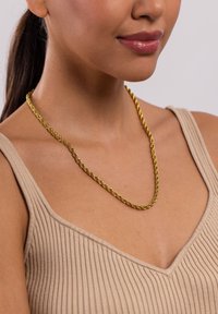 Eine goldene, verdrehte Kettenhalskette, die über einem gerippten beigen Oberteil getragen wird. Die Halskette hat eine geflochtene Textur und eine glatte Oberfläche.