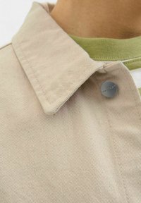 Beige corduroy fabric with a pointed collar, featuring a metal button on the left side. Underneath is a green ribbed crewneck.