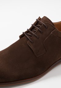 Scarpa da uomo in suede marrone con una superficie liscia, punta arrotondata, sei occhielli con lacci scuri e suola in pelle a contrasto.