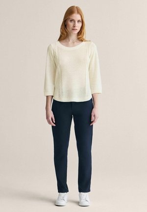 Rødhåret kvinde står iført en cremefarvet striktrøje, mørkeblå bukser og hvide sneakers mod en ensfarvet beige baggrund.