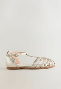 Metallic silver sandal with braided straps, T-bar design, and soft pink inner lining; features a buckle closure and flat sole.