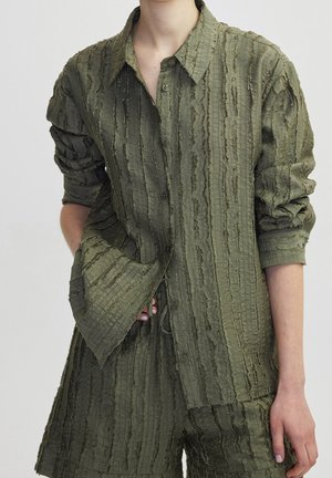 Kobieta nosi teksturowaną oliwkowo-zieloną koszulę z długim rękawem zapinaną na guziki oraz pasujące do niej szorty, ręka wsunięta w kieszeń koszuli, na jednolitym tle.