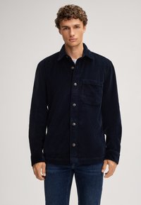 Camicia di velluto a corda blu navy con bottoni sul davanti, tasca sul petto e maniche lunghe. Presenta una texture a righe verticali e colletto standard.