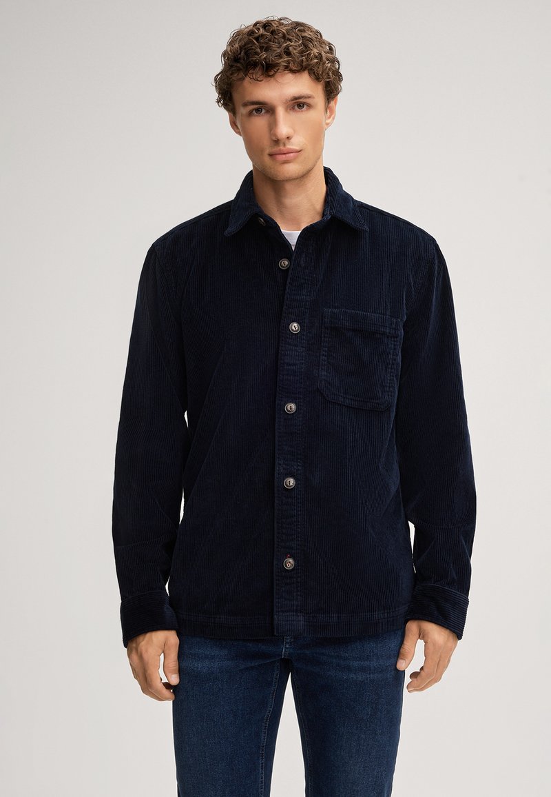 Camicia di velluto a corda blu navy con bottoni sul davanti, tasca sul petto e maniche lunghe. Presenta una texture a righe verticali e colletto standard.