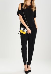 Sort kold- skulder top, sorte skræddersyede bukser og sorte hæle. Lille crossbody taske med gule, hvide og marineblå sektioner. Enkel kædestrop.