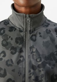 Grå treningsjakke med leopardmønster. Har høy krage, glidelås-lukking og strukturert stoff. Adidas-logo på siden.