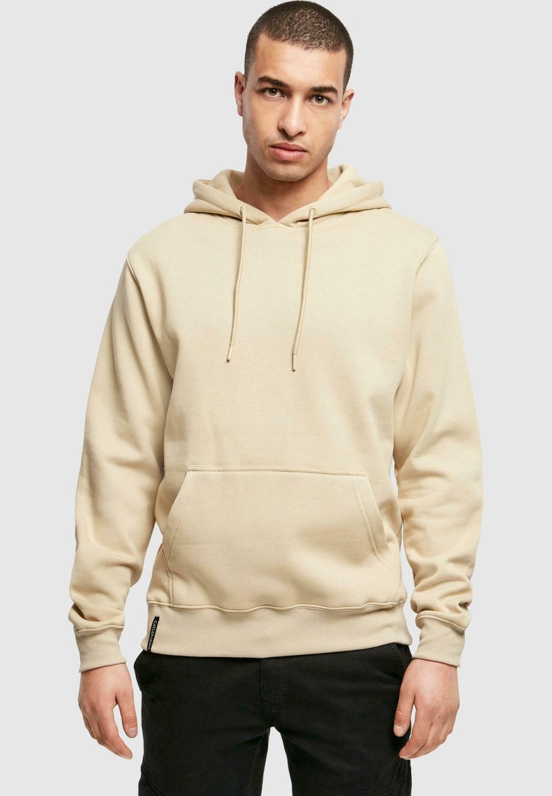 Beżowy hoodie z kieszenią kangurką, regulowanym sznurkiem, ściągaczami przy mankietach i u dołu, wykonany z miękkiego materiału, noszony na czarnych spodniach.