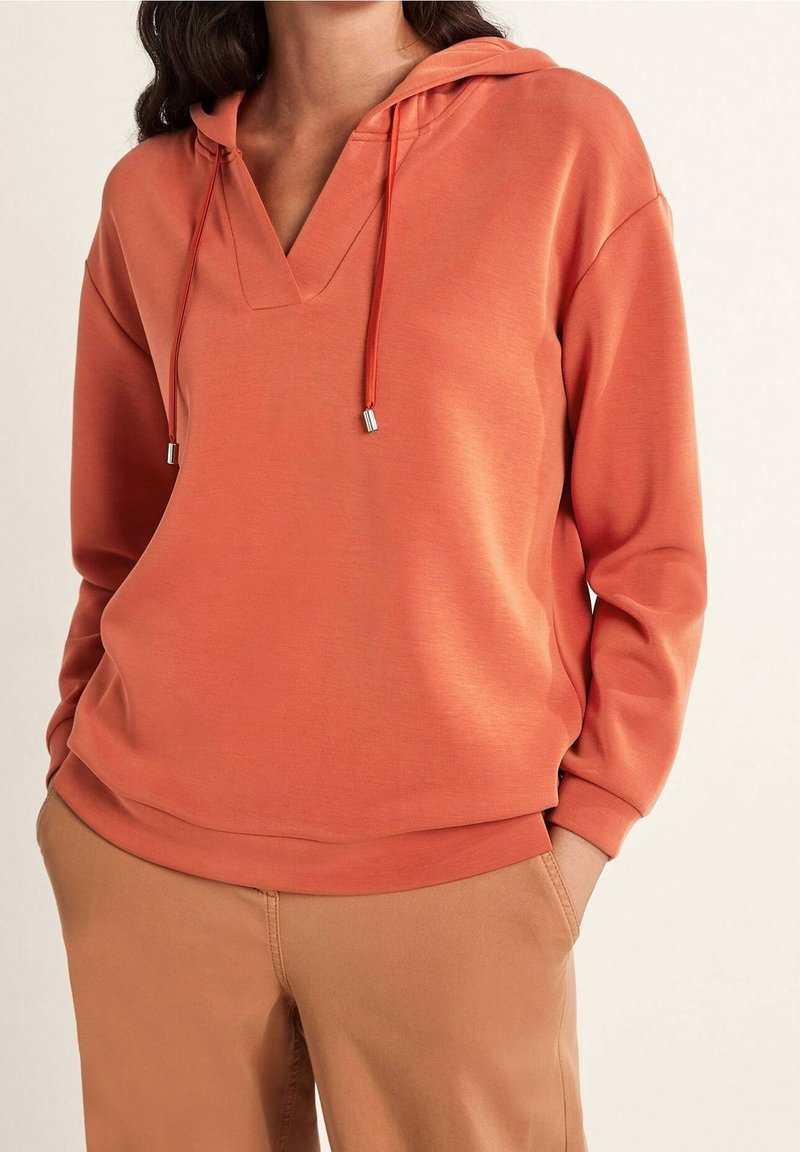 Femme portant un sweat à capuche orange ample à manches longues avec des cordons et un pantalon beige, les mains dans les poches.