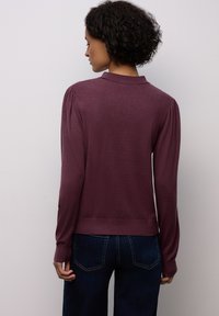 Bordeauxfarbener Pullover mit Kragen, langen Ärmeln und gerafftem Schulterdetail. Weicher Stoff, tailliert, kombiniert mit dunklen Jeans.
