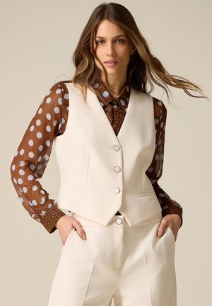 Vrouw draagt een crèmekleurig vest en broek, met een doorschijnende blouse met bruine stippen, handen in de zakken, los haar en een neutrale achtergrond.