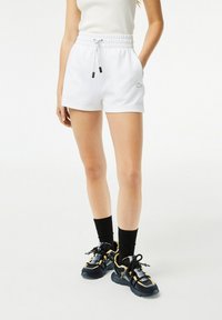 Pantaloni sportivi bianchi con vita elasticizzata, cordoncino e tasche laterali, abbinati a calzini neri e scarpe da ginnastica con accenti dorati.