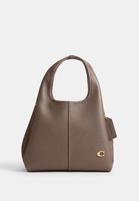 Sac à main en cuir grainé marron avec une poignée supérieure, un emblème de logo doré et une étiquette en cuir embossée sur fond blanc.