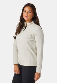 Lys beige fleece pullover med halv-lynlås krave, sidesømme og broderet logo. Glat tekstur og afslappet pasform. Sorte bukser er synlige.