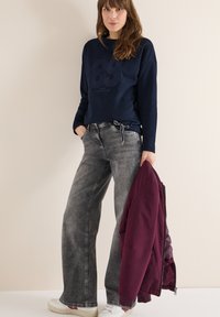 Marineblauer Pullover mit grafischem Aufdruck, graue Jeans mit weitem Bein und eine dunkelrote Bomberjacke. Strukturierter Stoff, lässige Passform und Sneaker in neutralen Tönen.