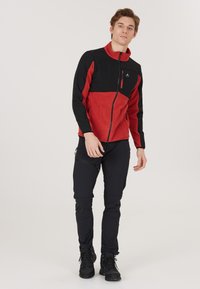 Röd och svart zip-up fleecejacka med hög krage och bröstficka, kombinerad med svarta byxor och mörka skor, lämplig för utomhusaktiviteter.