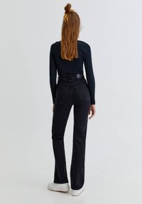 Jean noir évasé taille haute avec une texture lisse, poches arrière et une ceinture étroite, associé à des baskets blanches.