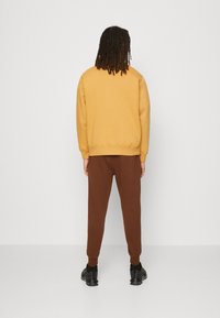 Gul sweatshirt med avslappnad passform, tillsammans med bruna avsmalnande joggingbyxor och svarta sneakers, sett från baksidan. Slät textur.