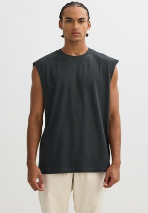 Joven con cabello trenzado, vestido con una camiseta sin mangas gris oscuro y pantalones beige claros, frente a un fondo liso.