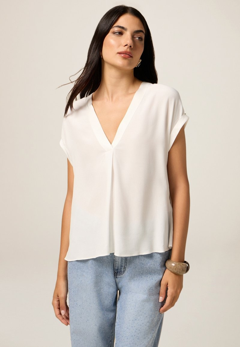 Blusa blanca de manga corta con escote en V, fabricada en tejido ligero, que presenta un corte holgado y una textura suave. Llevada con jeans azules.