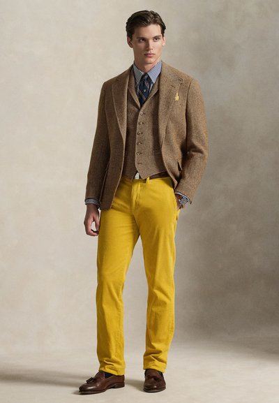 Brązowa marynarka z tweedu w jodełkę, nałożona na jasnoniebieską koszulę w paski. Żółte spodnie sztruksowe i ciemnobrązowe buty.