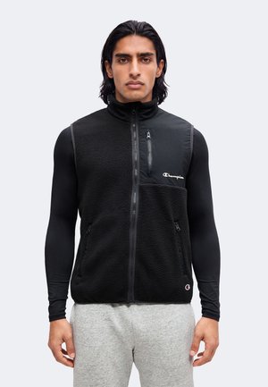 Man draagt zwart fleecevest met ritssluiting over zwarte lange mouwen en lichtgrijze joggingbroek, staand tegen een effen lichte achtergrond.
