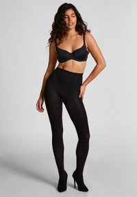 Schwarze undurchsichtige Strumpfhosen mit hohem Bund, kombiniert mit einem schwarzen BH. Das Modell posiert in High Heels und zeigt die Passform und Textur des Kleidungsstücks.